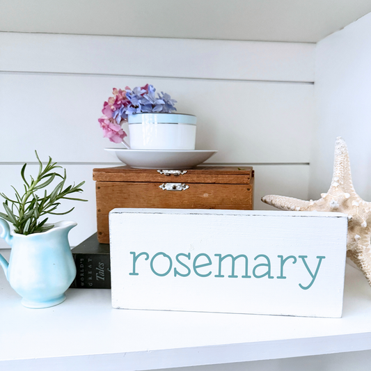 White sign labeled 'rosemary' on a shelf with decorative items including a starfish and a rosemary plant.