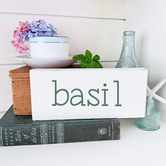 Decorative setup with a sign labeled 'basil', books, and a small plant on a white surface.