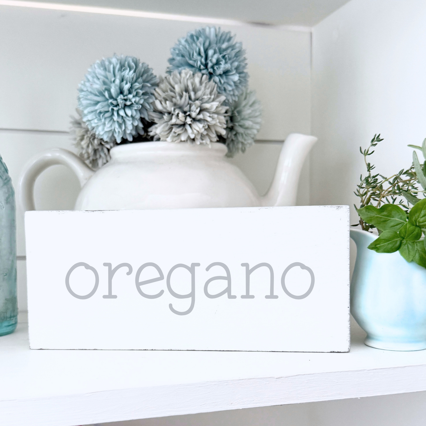 White teapot with decorative elements on a shelf with a 'oregano' sign.