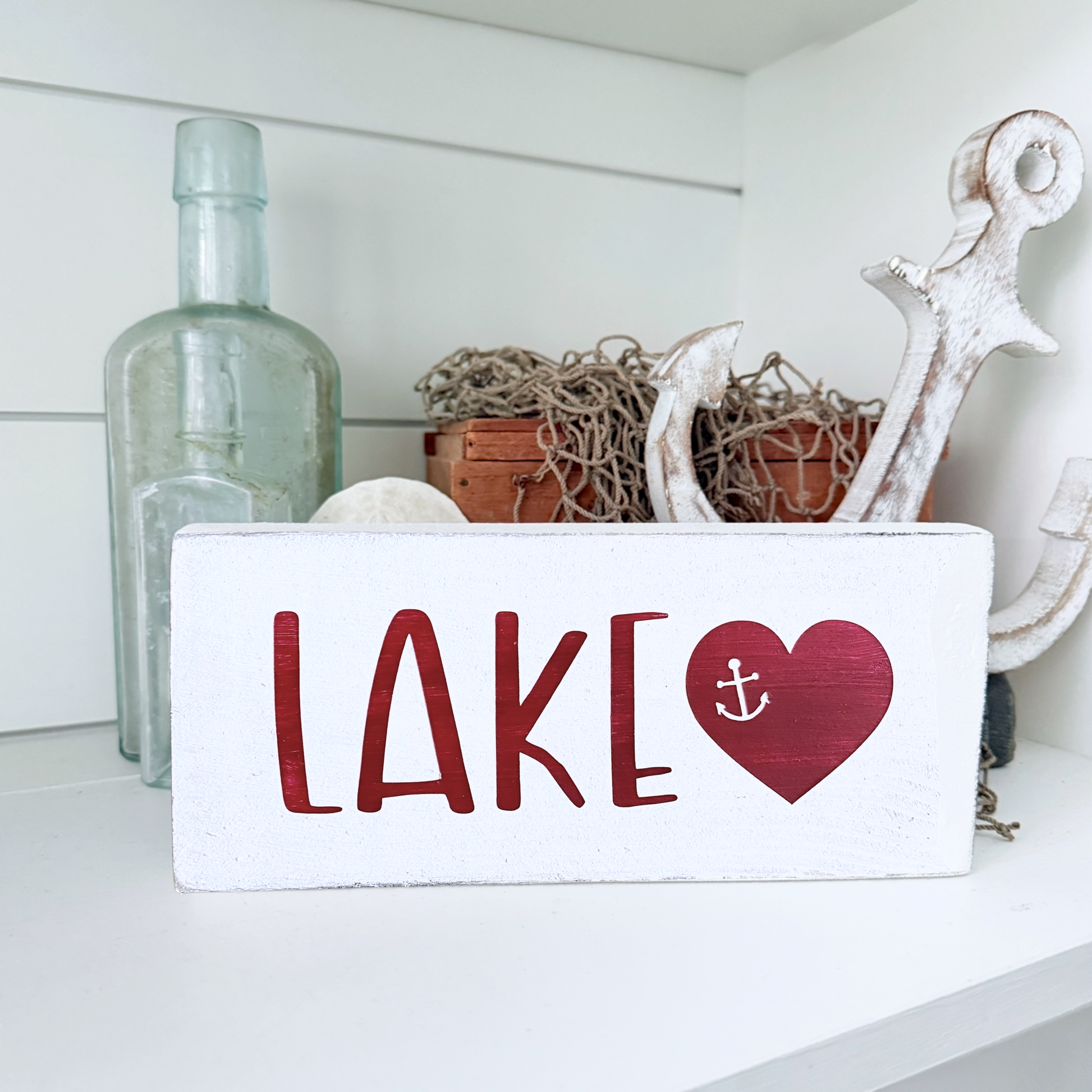 A white rectangular wooden sign with the word "LAKE" in red letters and a red heart containing a small white anchor, displayed on a white shelf with nautical decor props including a large white anchor, a glass bottle, and rope.
