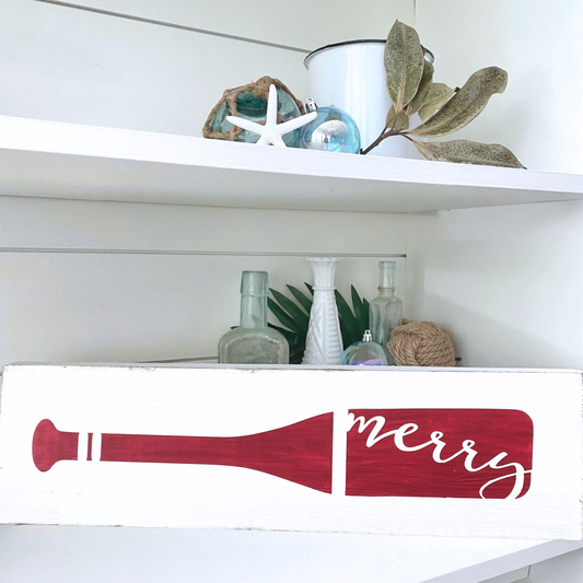 A white wooden sign with a red painted paddle stroke and the word 'merry' in white script, styled on a white shelf with nautical and home decor items.