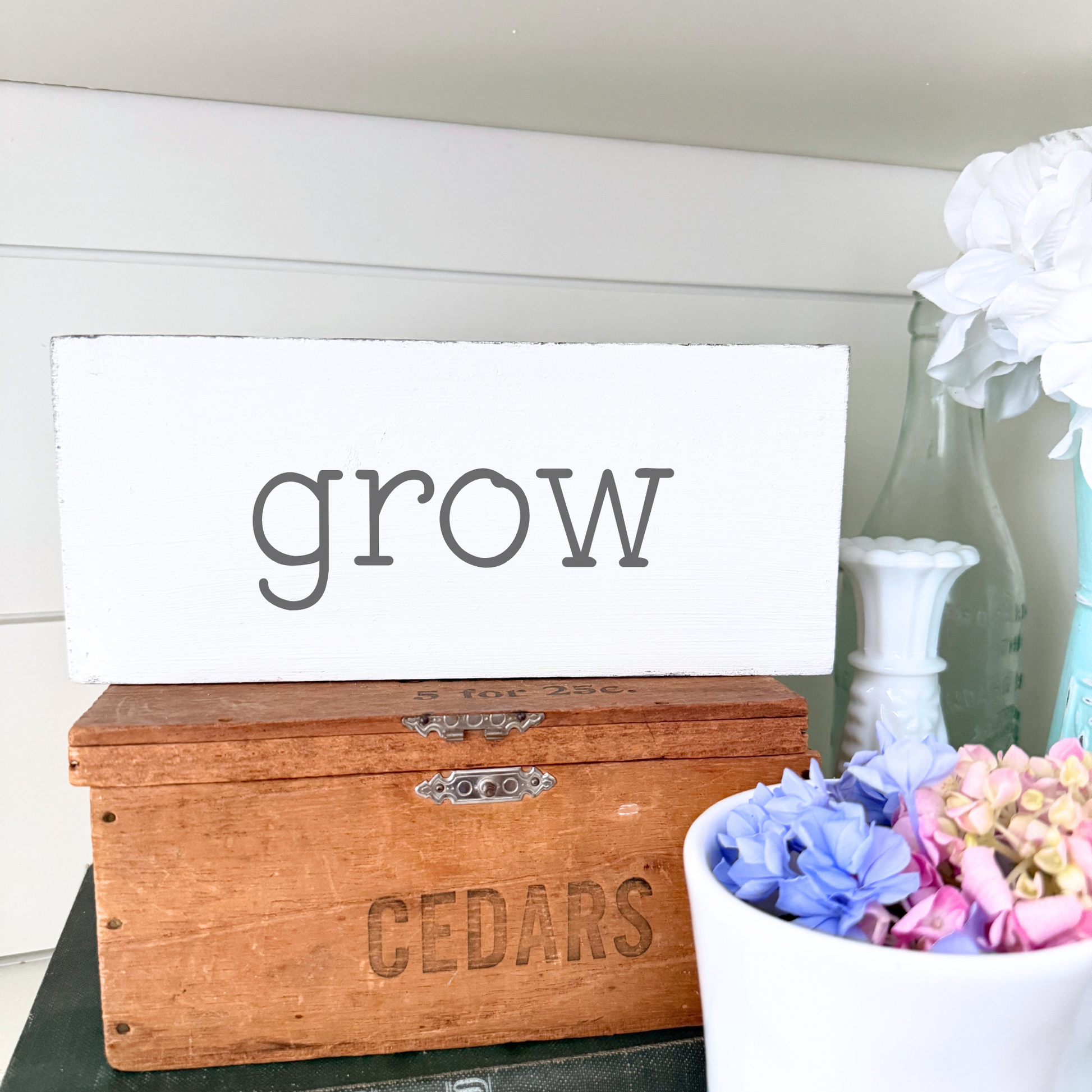 Wooden box with 'CEDARS' engraving, white sign with 'grow' text, and flowers in a vase.