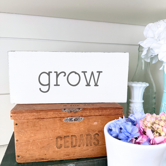 Wooden box with 'CEDARS' engraving, white sign with 'grow' text, and flowers in a vase.