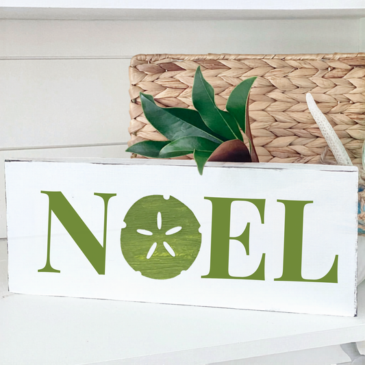 A white rectangular decorative sign with the word "NOEL" in olive green, where the letter O is replaced by a sand dollar motif. The sign is styled on a white shelf with a woven basket, a green plant, and a white starfish behind it.