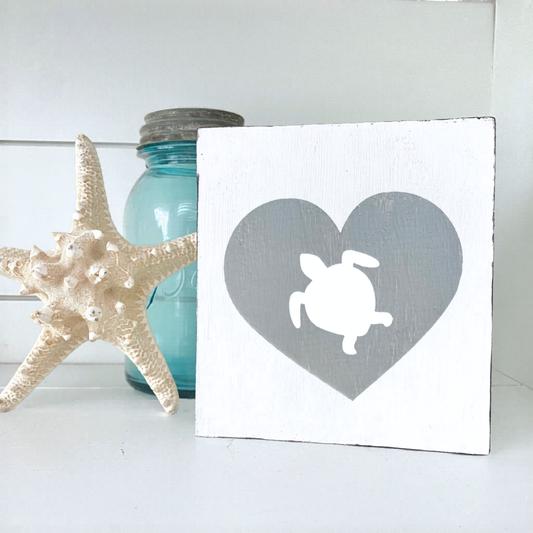 A square white wooden sign with a gray heart and white turtle silhouette, styled on a white surface next to a starfish and a blue glass jar.