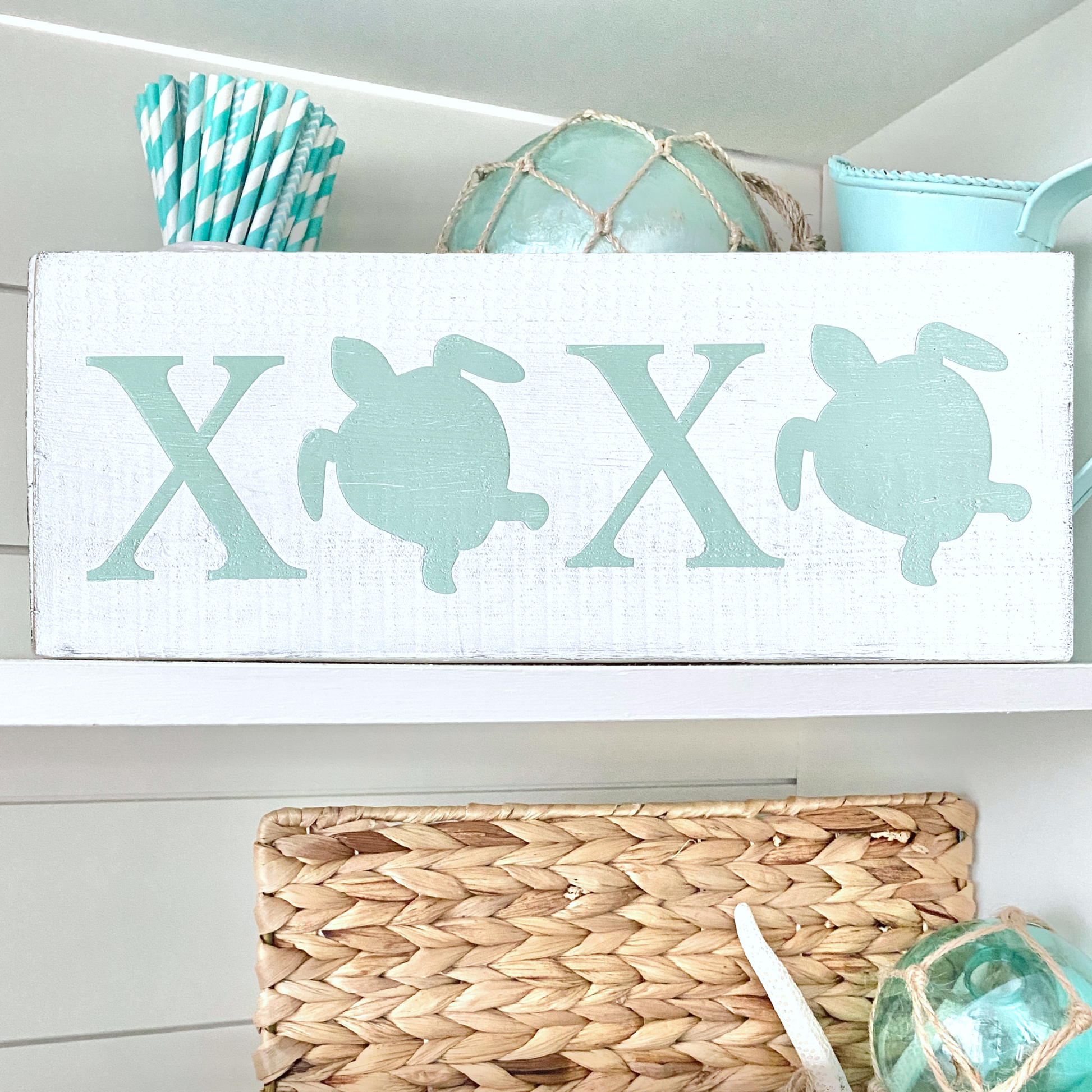 A rectangular white wooden sign with two large mint-green letters X and two matching turtle silhouettes to express XOXO in a coastal valentine way,  displayed on a white shelf with beach-themed decor.