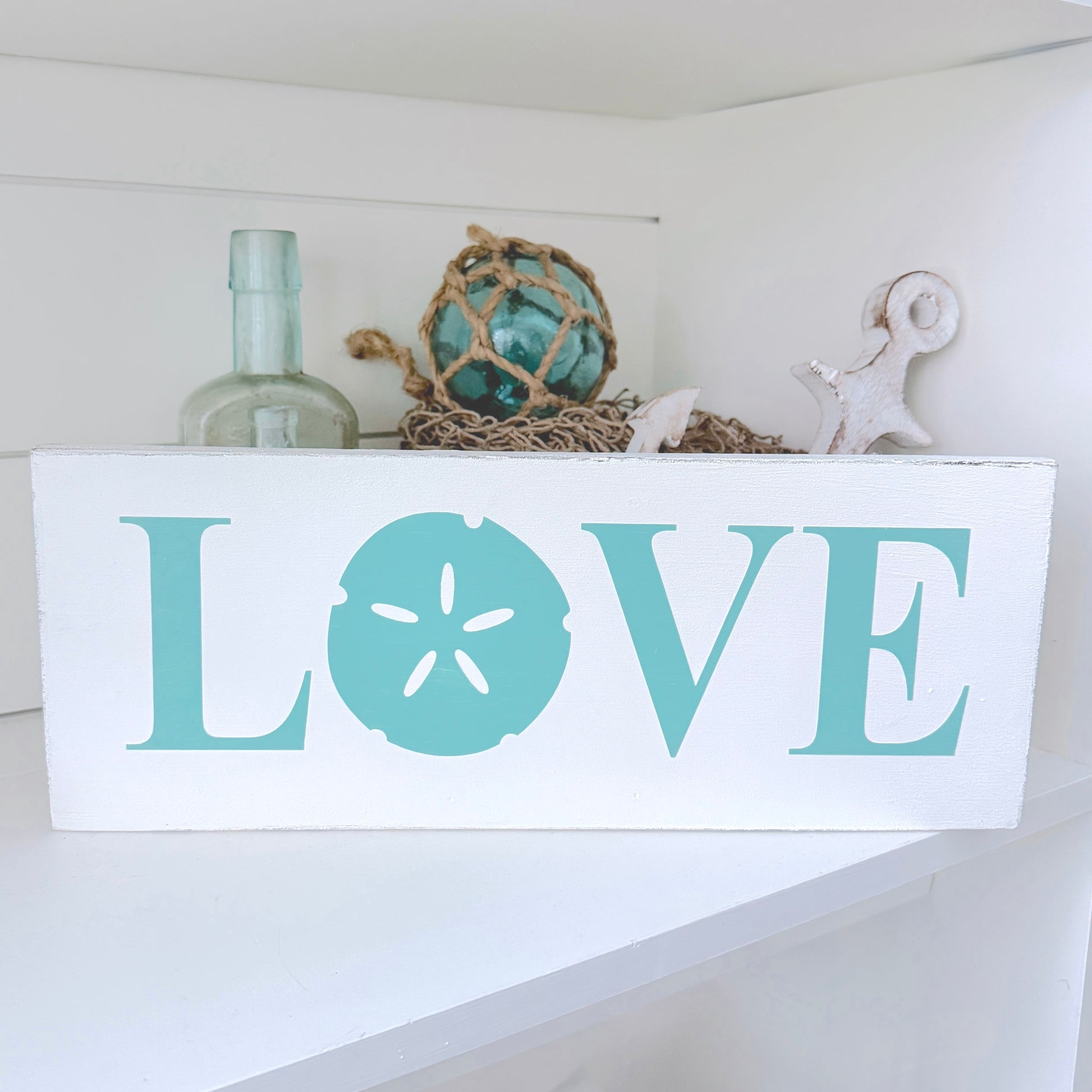 A white rectangular wooden sign with the word LOVE in large aqua letters, where the O is shaped like a sand dollar. The sign sits on a white shelf with nautical decor behind it, including a glass bottle, rope ball, starfish, and shell.