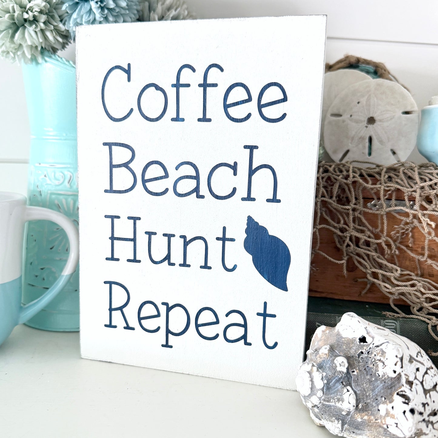 Sign with 'Coffee Beach Hunt Repeat' text on a decorative background with mugs and shells.