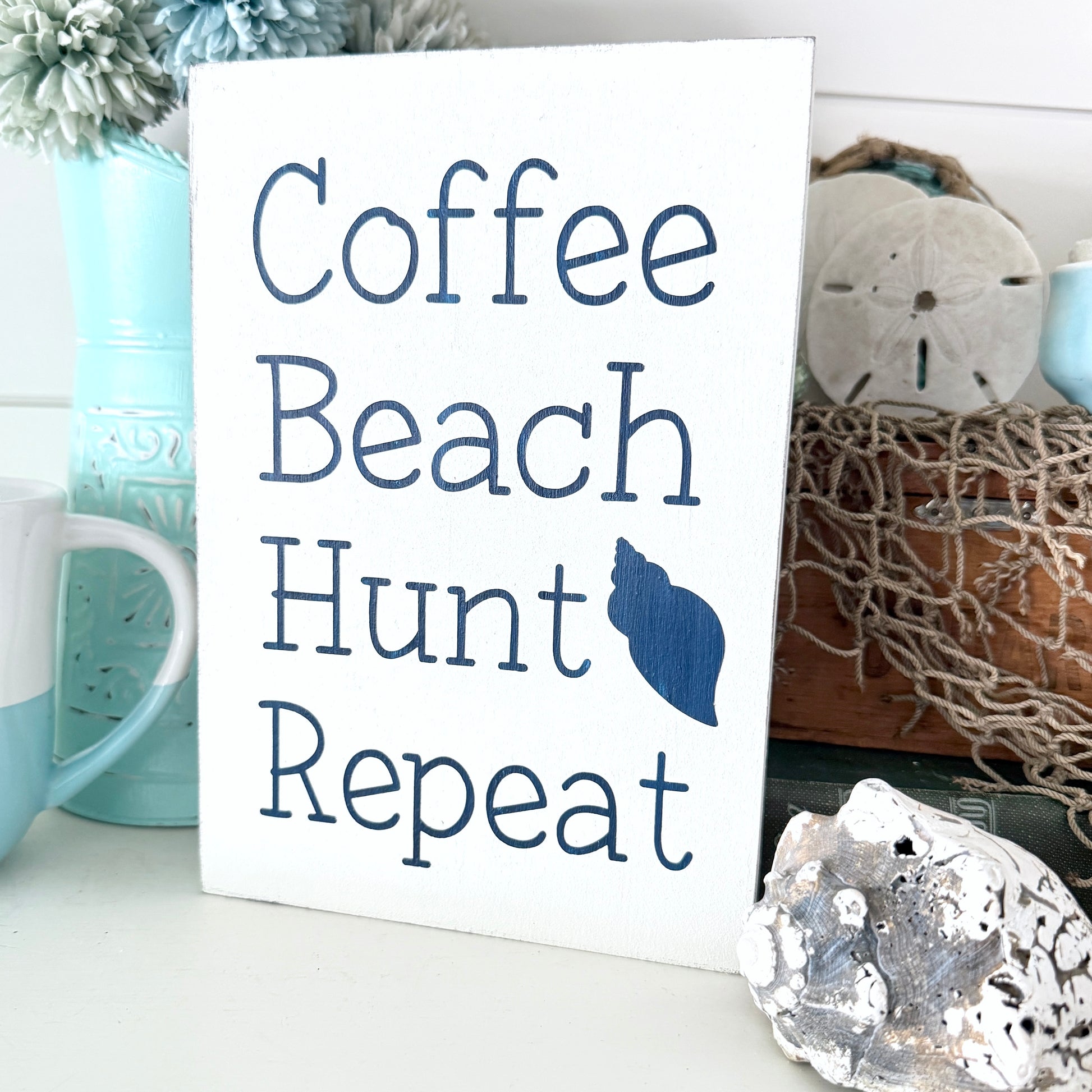 Sign with 'Coffee Beach Hunt Repeat' text on a decorative background with mugs and shells.