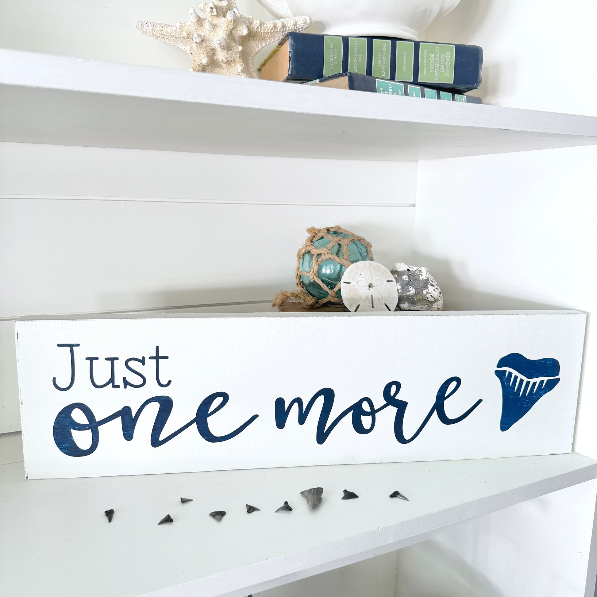 Decorative sign with 'Just one more' text on a white shelf with decorative items.