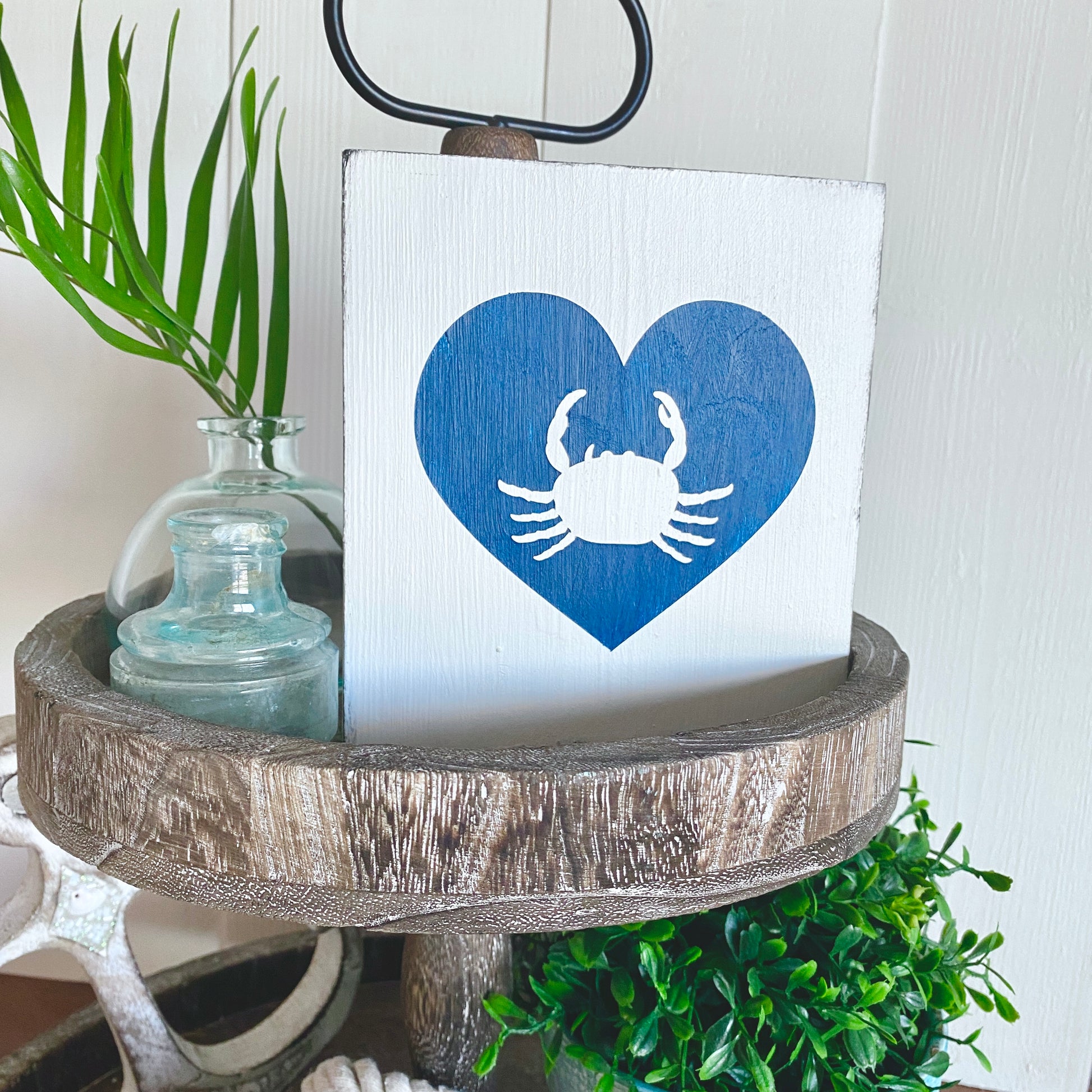 Decorative sign with a blue heart and white crab design on a wooden stand with plants.