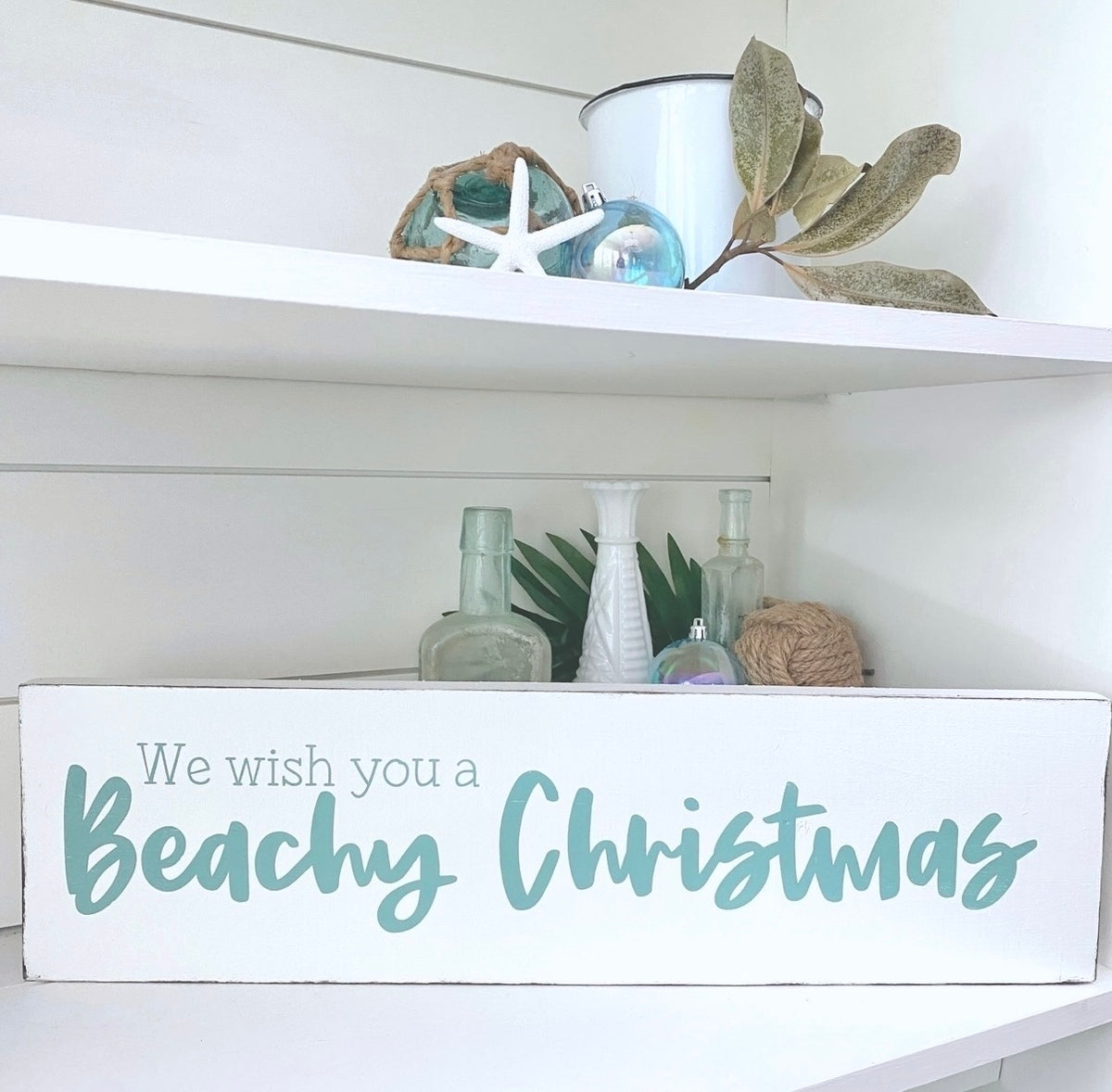 A white rectangular decorative sign on a shelf that reads 'We wish you a Beachy Christmas' in aqua script and serif text. The sign is styled on a white shelf with beach-themed decor, including a white bucket, starfish, green leaves, and glass bottles.