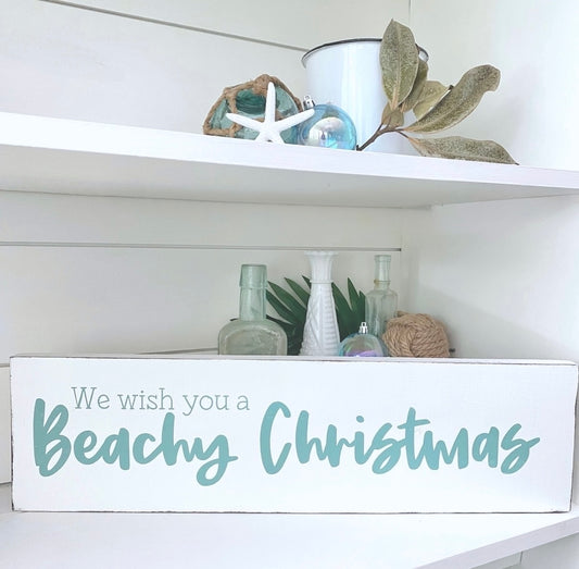 A white rectangular decorative sign on a shelf that reads 'We wish you a Beachy Christmas' in aqua script and serif text. The sign is styled on a white shelf with beach-themed decor, including a white bucket, starfish, green leaves, and glass bottles.