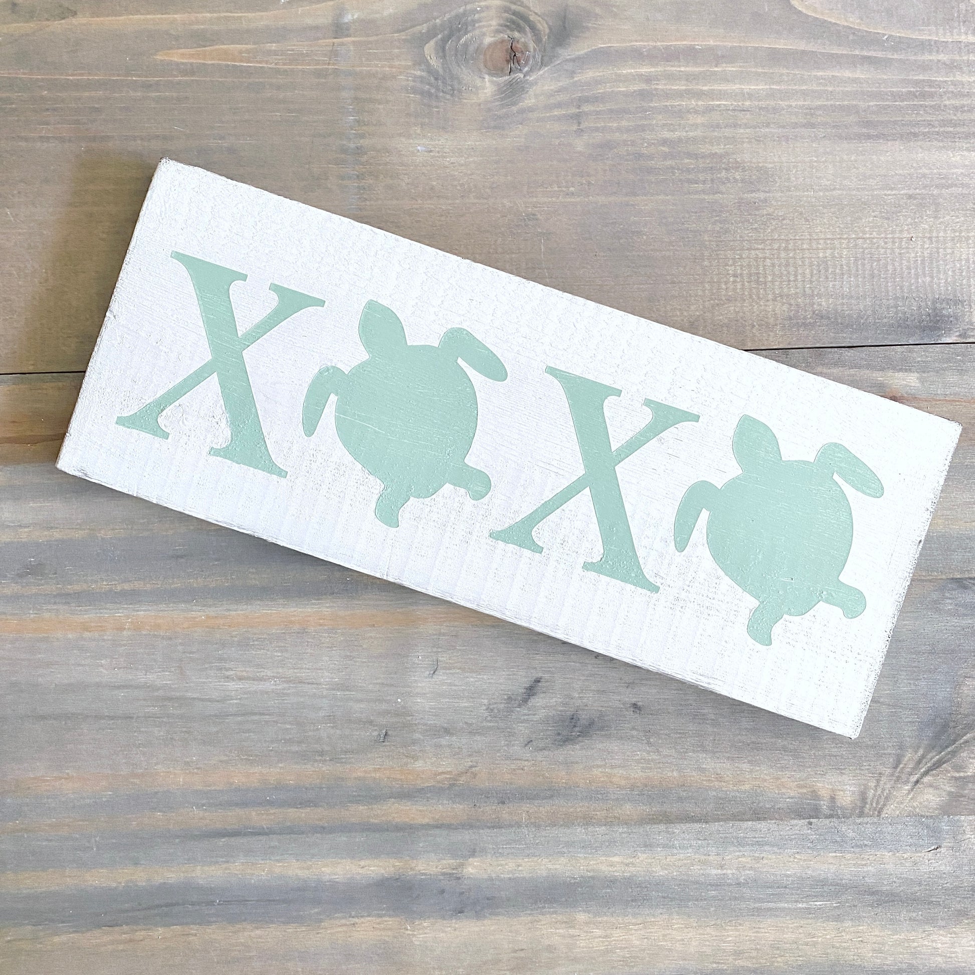 A rectangular white wooden sign with a distressed finish, featuring the letters 'X' and 'O' with sea turtle silhouettes in a mint color. The sign is photographed at an angle on a rustic, weathered gray-brown wooden surface.