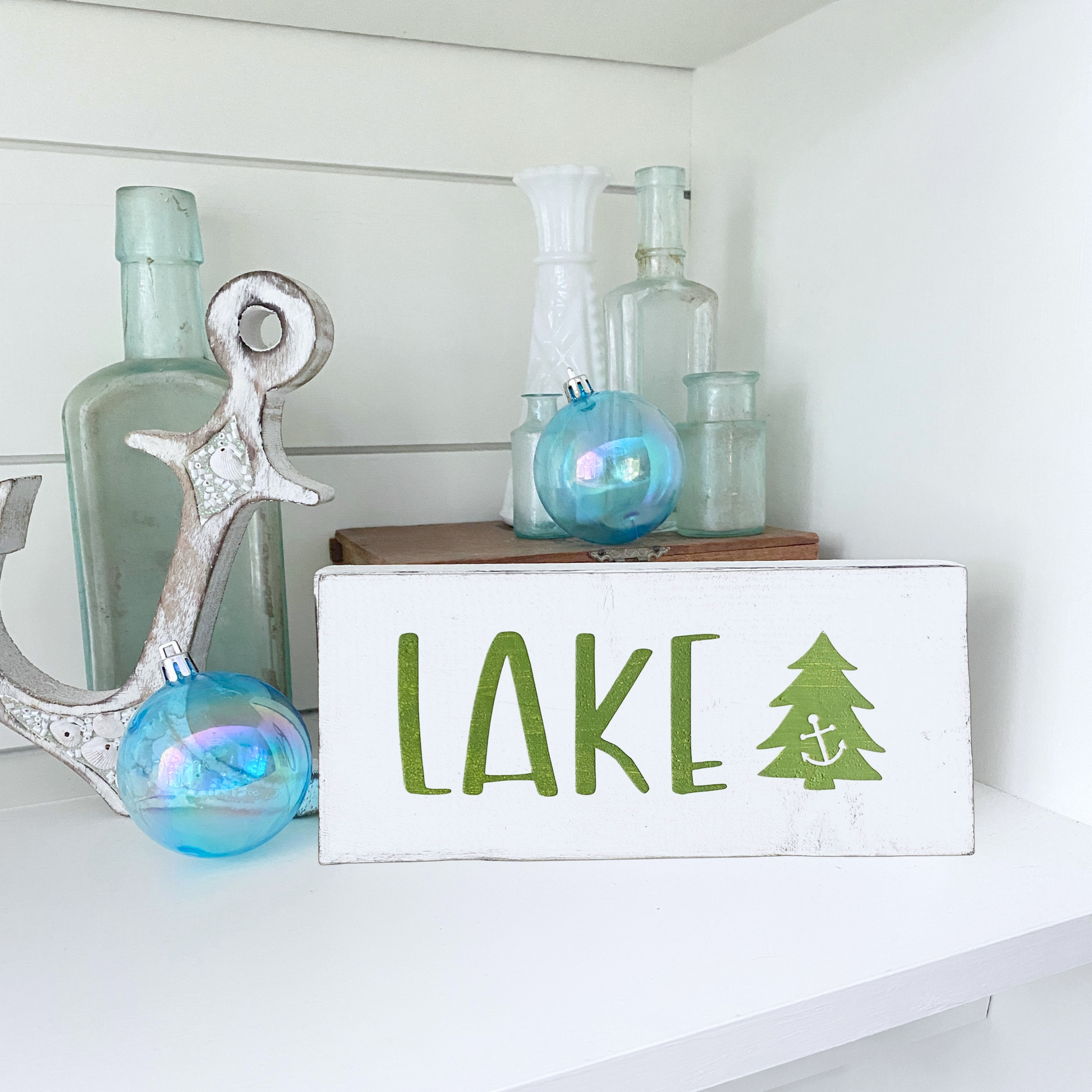 A white rectangular decorative sign with the word "LAKE" in green and a green tree with a small anchor inside, styled on a white shelf with glass bottles and blue ornaments.