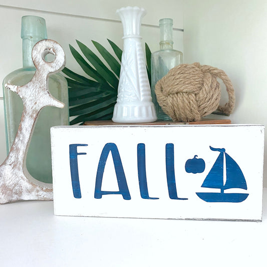 Decorative sign with 'FALL' and sailboat design, surrounded by decorative items on a white surface.