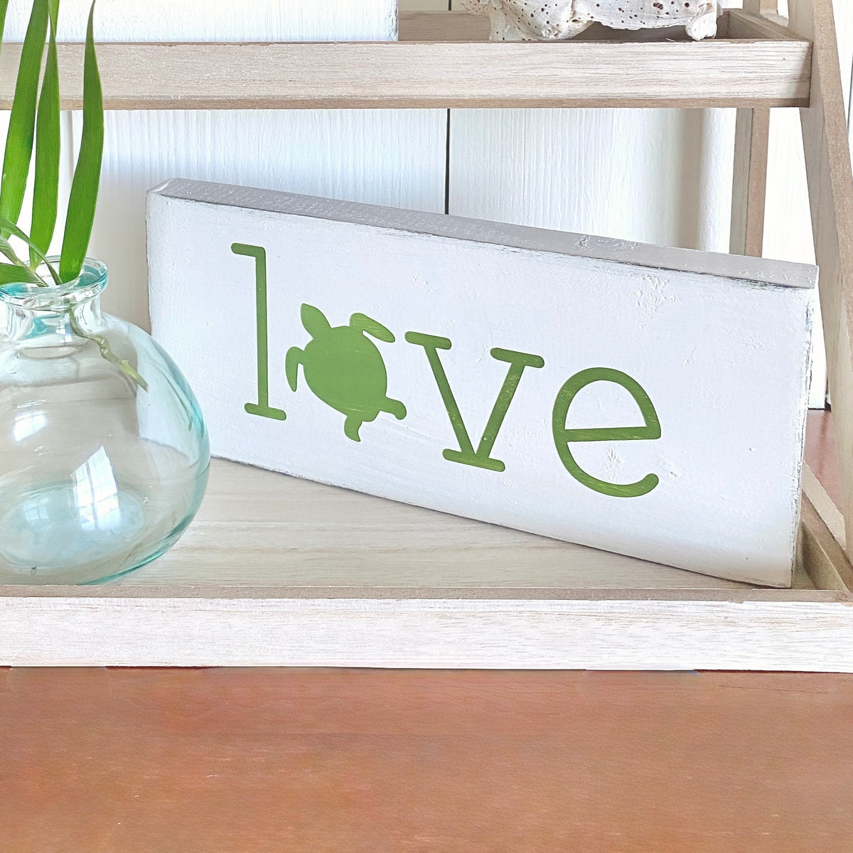 Decorative sign with 'love' and sea turtle design on a wooden shelf, displayed by a vase and palm frond