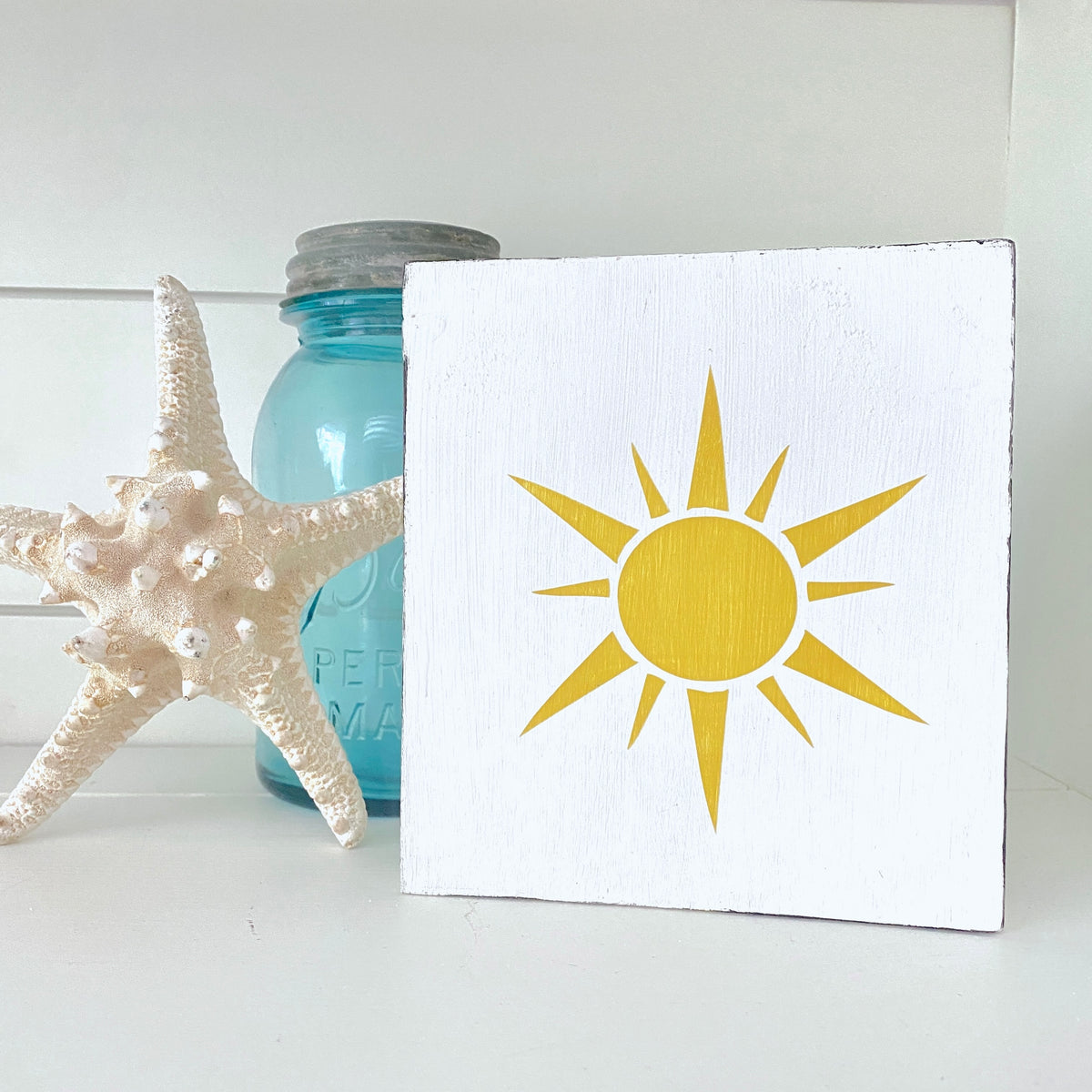 Sunshine sign, white sign with yellow sun graphic displayed on a shelf with a starfish and vintage blue jar