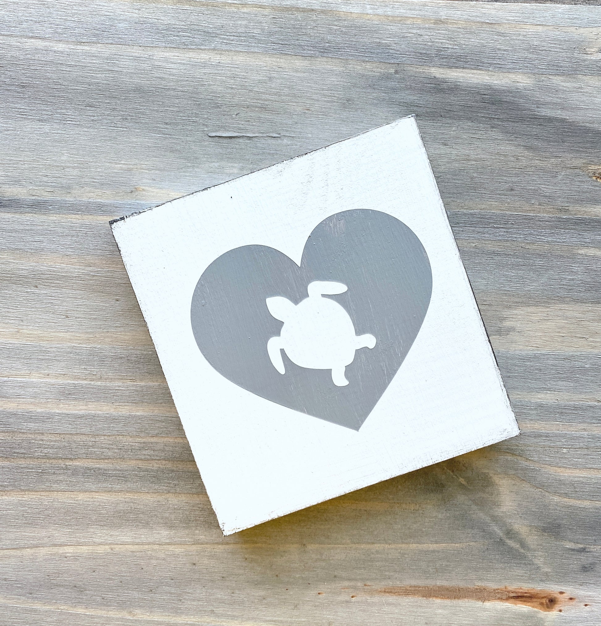 A square white wooden sign with a gray heart design containing a white sea turtle silhouette, placed on a rustic gray wood surface.