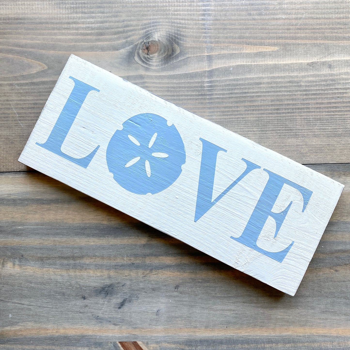 A rectangular white wooden sign with the word LOVE, where the O is replaced by a light blue sand dollar graphic, placed diagonally on a rustic dark wood plank surface.