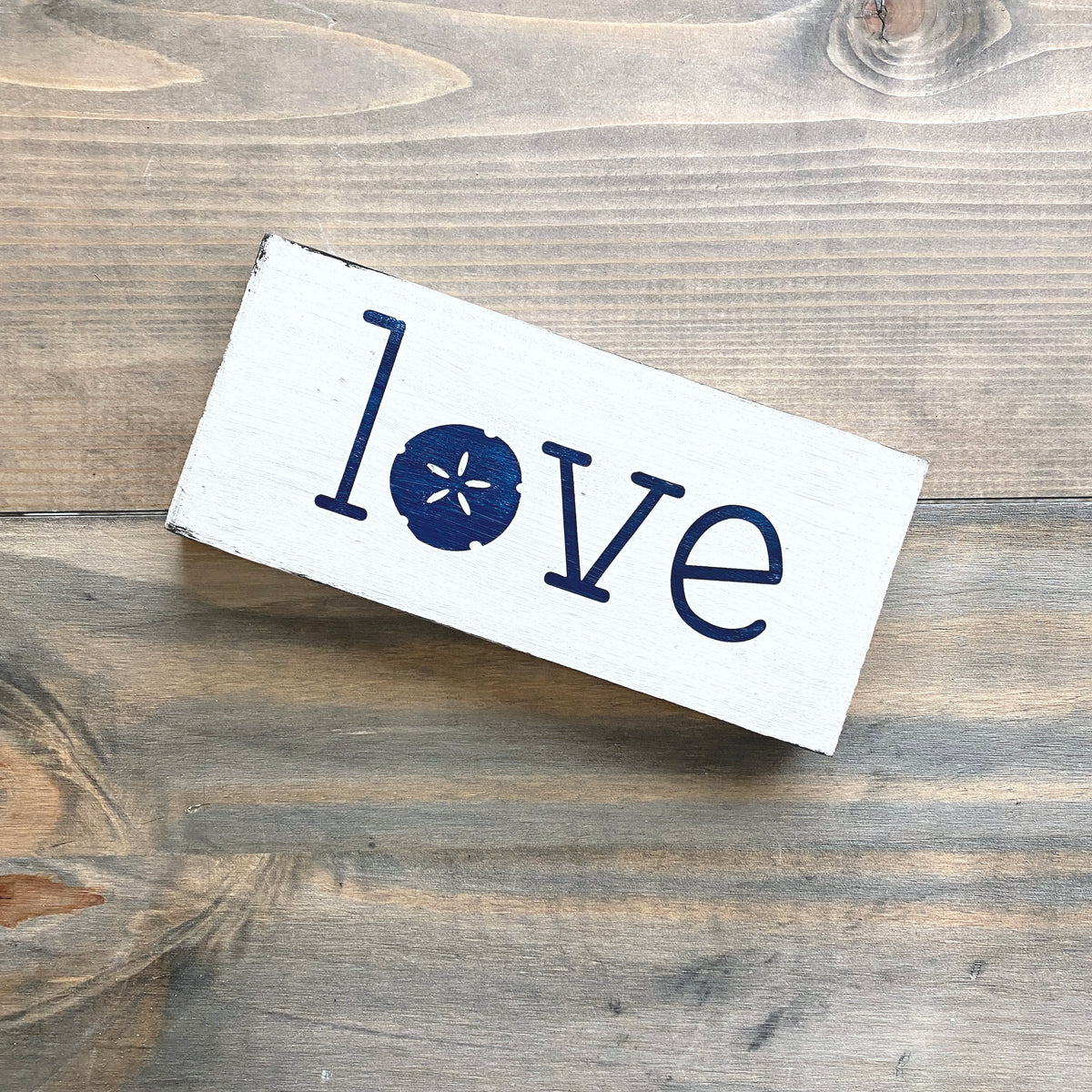 A rectangular white wooden sign with the word 'love' painted in navy blue, where the letter 'o' is replaced by a navy sand dollar motif. The sign is photographed from above on a rustic, weathered gray-brown wooden surface.