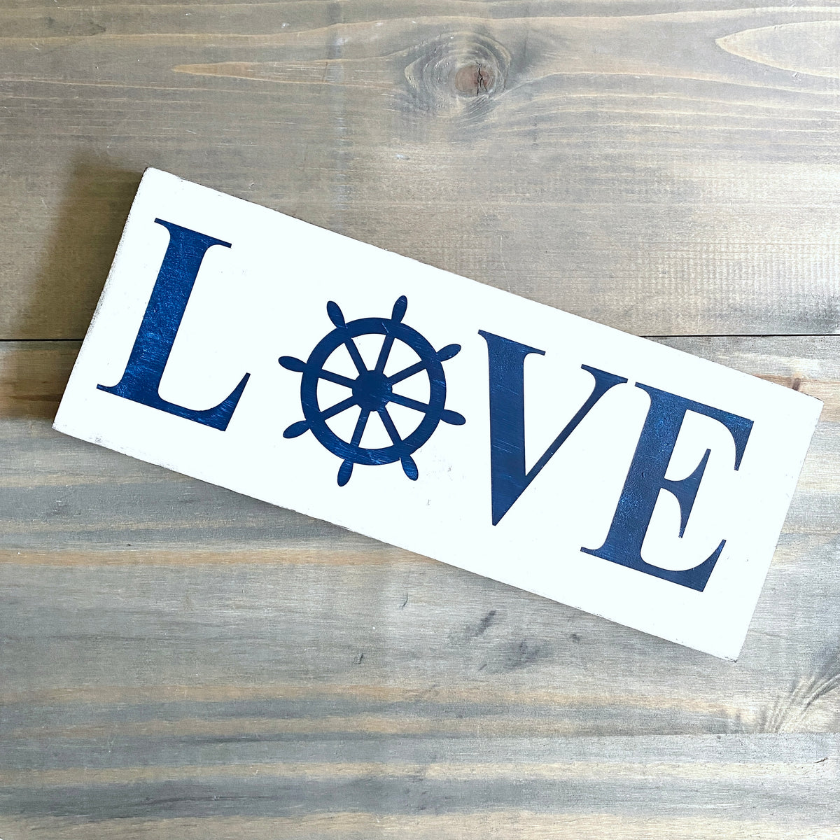 A rectangular white wooden sign with the word LOVE, where the O is replaced by a navy blue ship's wheel, displayed diagonally on a rustic gray wood plank surface.