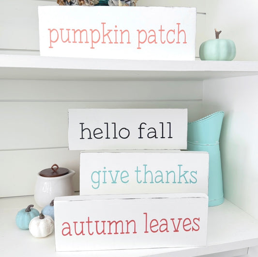 Stack of autumn-themed wooden signs on a shelf with decorative pumpkins.