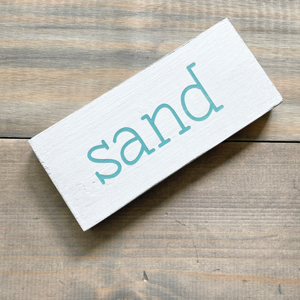 A rectangular white wooden sign with the word 'sand' painted in a light teal lowercase font, shown on a rustic brown wood plank surface.