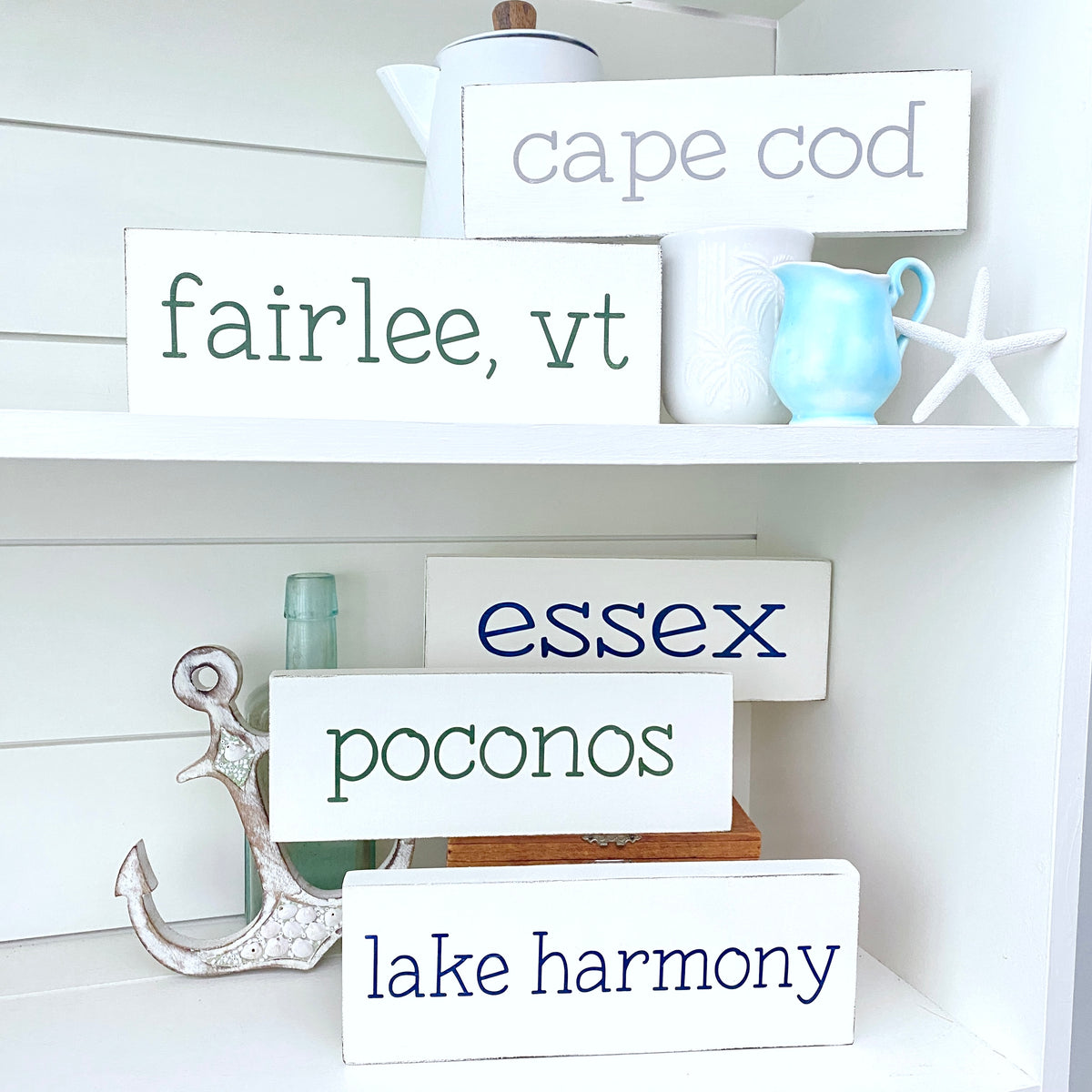 A set of white rectangular decorative signs with coastal-themed phrases including 'cape cod', 'fairlee, vt', 'essex', 'poconos', and 'lake harmony', styled on white shelves with nautical props like an anchor, a light blue pitcher, a white starfish, and ceramic mugs.