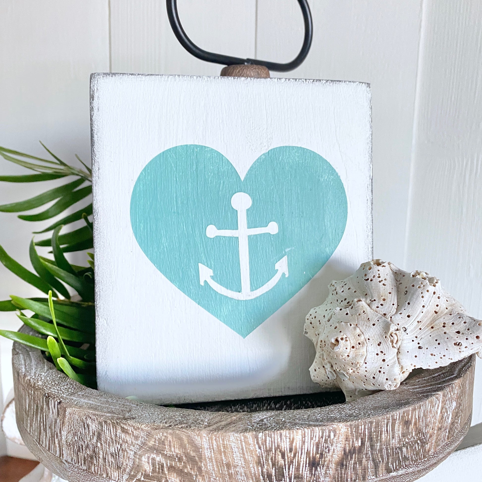 Decorative sign with a heart and anchor design on a white background, placed in a wooden bowl with a plant and shell.